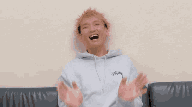 a young man in a hoodie is sitting on a couch with his mouth open and his hands in the air .