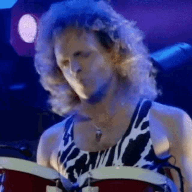 a man with long curly hair is playing drums on a stage in a cow print tank top .