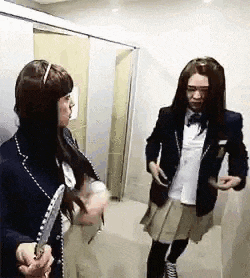 two women in school uniforms are standing next to each other in a hallway . one of the women is holding a knife .