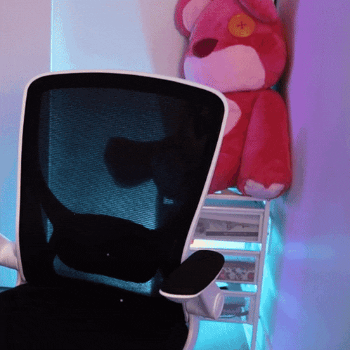 a pink teddy bear with a yellow button on its face sits behind a black chair