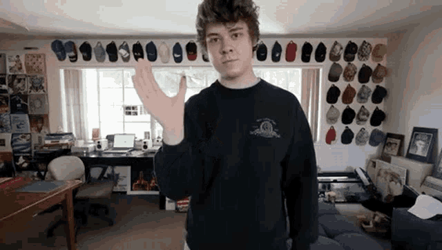a young man wearing a black sweater is standing in a living room with many hats on the wall .