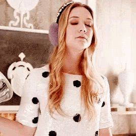a woman wearing ear muffs and a white dress with black polka dots is standing in front of a blackboard .