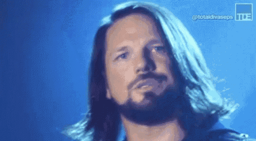 a man with long hair and a beard is standing in front of a blue background and looking at the camera .