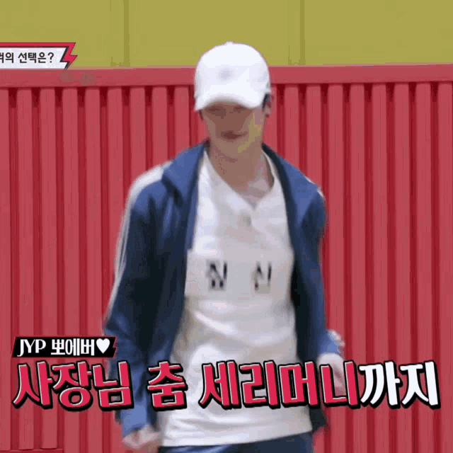 a man wearing a white hat and a blue jacket stands in front of a red fence with chinese writing on it