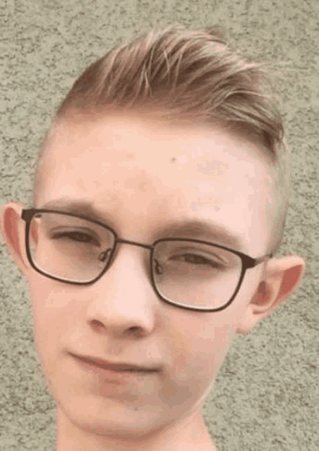 a young boy wearing glasses is standing in front of a wall and looking at the camera .