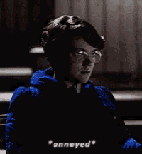 a young boy wearing glasses and a blue jacket is sitting in a dark room and looking annoyed .