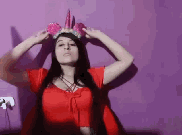 a woman wearing a red top and a pink unicorn crown is standing in front of a purple wall .