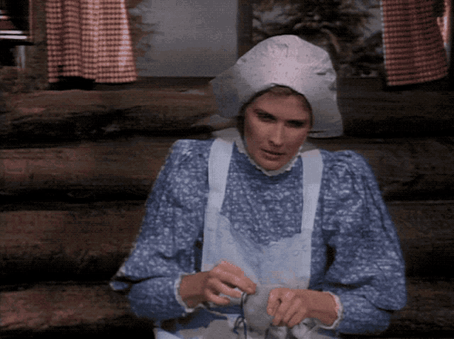 a woman in a blue floral dress and white apron is knitting