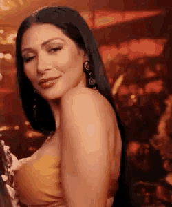a woman with long black hair is wearing a yellow top and earrings and looking at the camera .