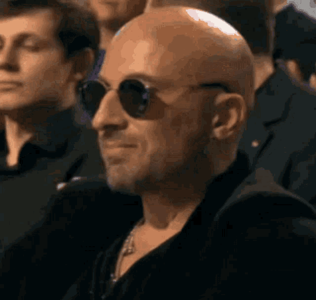 a bald man wearing sunglasses is smiling in a crowd of people .