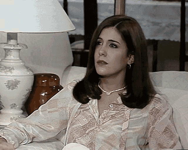 a woman wearing a pearl necklace sits on a couch next to a lamp