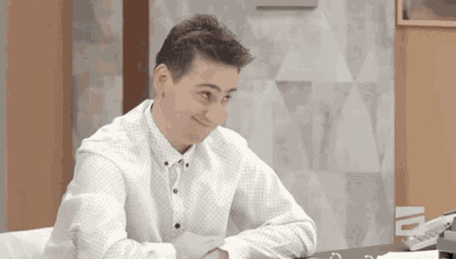 a man in a white shirt is sitting at a desk with his arms crossed and smiling .