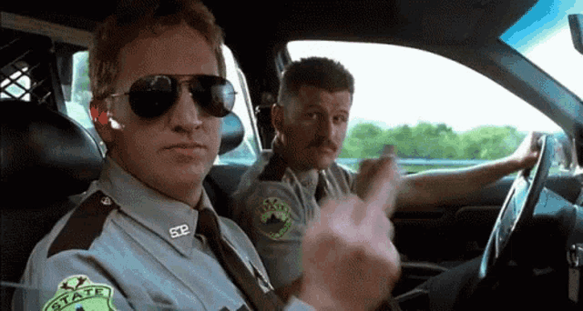 two sheriff 's officers are sitting in a car and one of them is giving the middle finger .