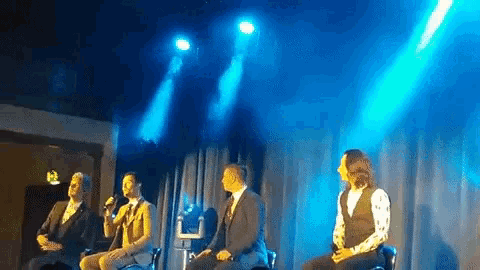 a group of men are sitting on a stage with microphones in front of a blue light .