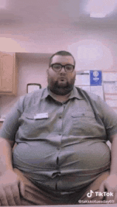 a man with a beard and glasses is sitting at a desk with his hands on his hips .