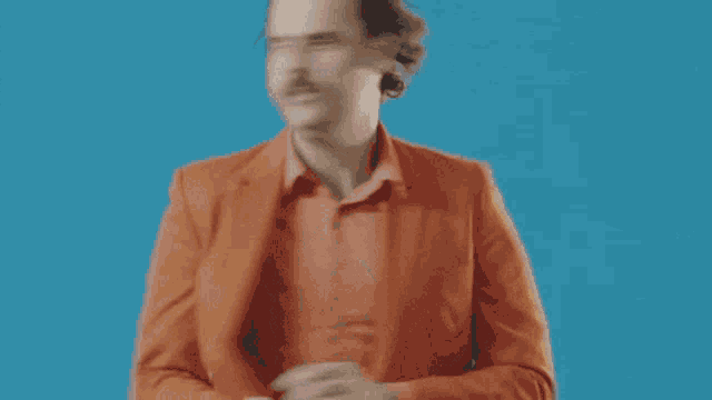 a bald man in an orange suit is standing in front of a blue background and smoking a cigarette .