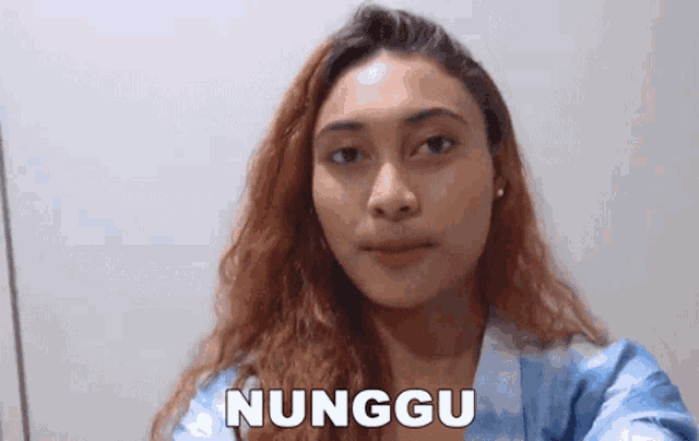 a woman in a blue robe is making a funny face and the word nunggu is on the bottom of her face .