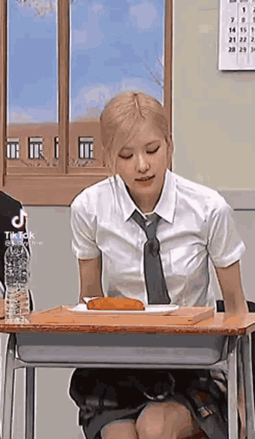 a girl in a school uniform and tie is sitting at a desk with a plate of food .