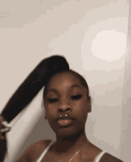 a woman in a white tank top is holding her hair in a ponytail and making a funny face .