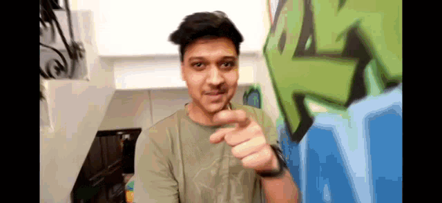 a man in a green shirt is pointing at the camera in front of a graffiti wall .