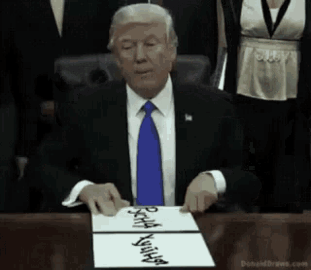 a man in a suit and tie is sitting at a table with a piece of paper that says " donald drews.com "