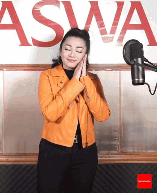 a woman in an orange jacket stands in front of a microphone in front of a wall that says aswa