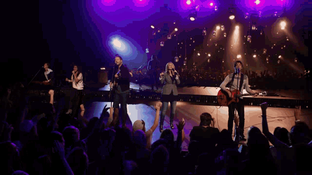 a group of people singing and playing guitars on a stage with purple lights