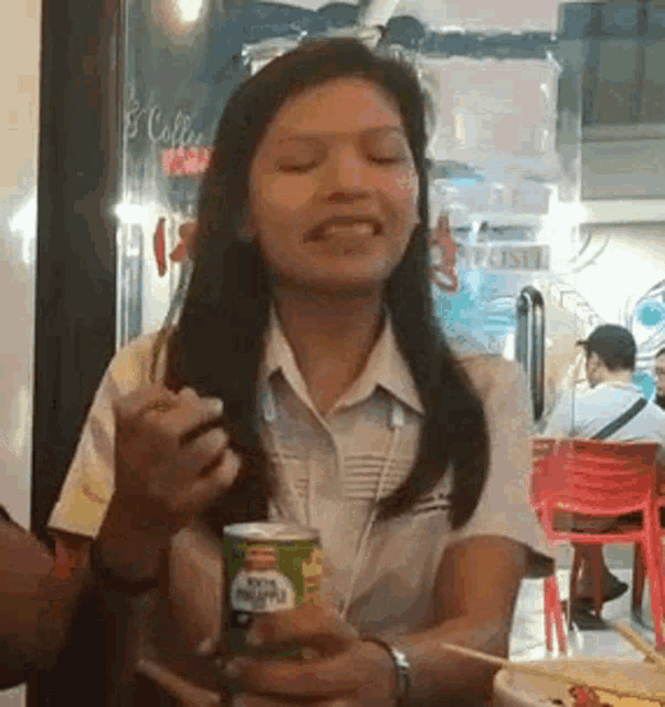 a woman is sitting at a table holding a can of soda and eating a sandwich .