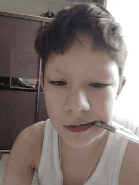 a young boy in a white tank top holds a green pen in his mouth