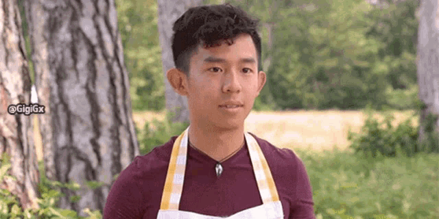 a young man wearing an apron is standing in front of trees .
