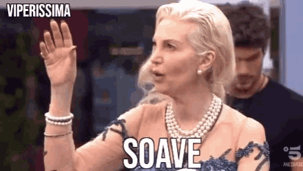 a woman wearing a pearl necklace and a blue dress is waving her hand and saying soave .
