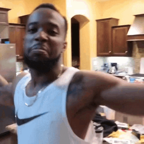 a man wearing a white nike tank top is standing in a kitchen with his arms outstretched .