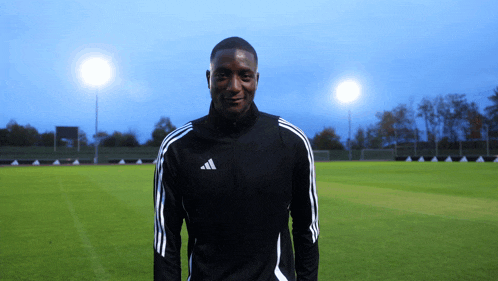 a man wearing an adidas jacket is standing on a soccer field
