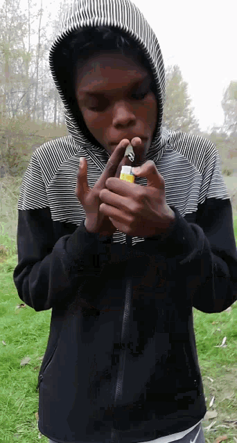 a young man in a black and white striped hoodie is lighting a cigarette