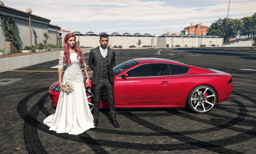 a bride and groom pose in front of a red sports car in a parking lot