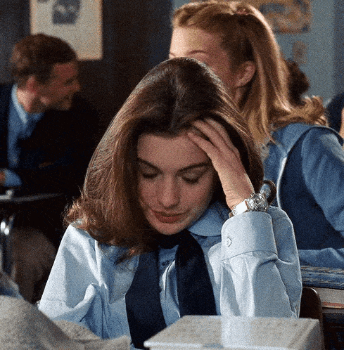 a girl in a blue shirt and tie is sitting at a desk with her hand on her forehead