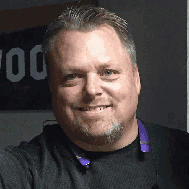 a man wearing a black shirt with a purple neckband smiles for the camera
