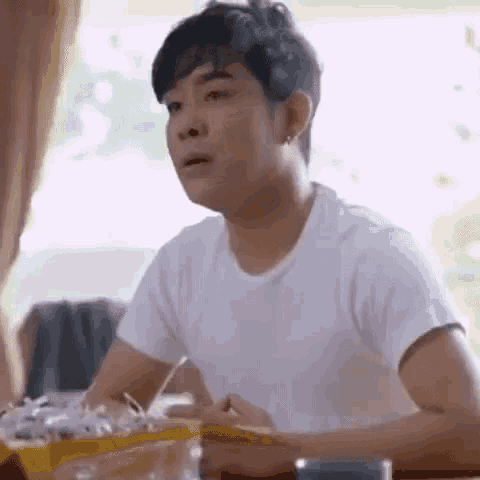a man in a white t-shirt is sitting at a table with chopsticks in his hand .