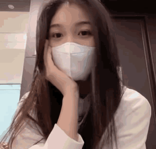 a woman wearing a white face mask rests her hand on her chin