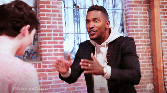 a man in a suit is talking to another man in front of a brick wall .