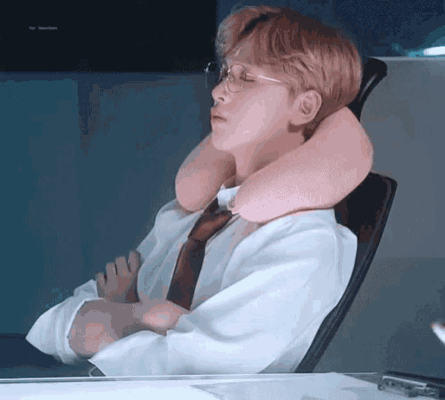 a man wearing glasses and a pink neck pillow is sitting in an office chair