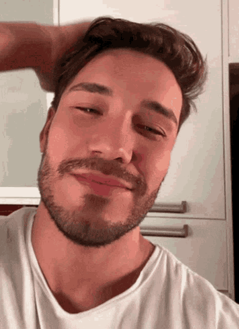 a man with a beard and a white shirt is smiling with his hand on his head
