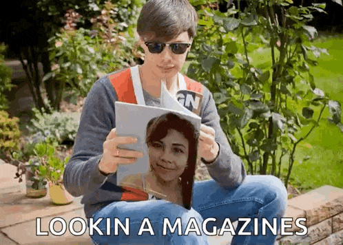 a young man wearing sunglasses is reading a magazine with a picture of a woman on the cover .