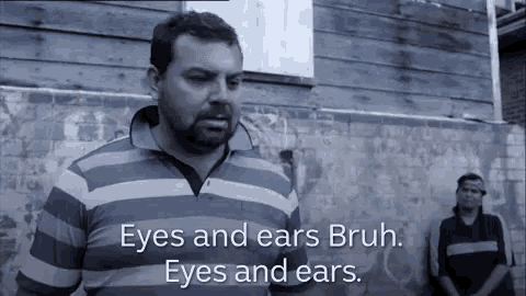 a man in a striped shirt is standing in front of a wooden building and talking about his eyes and ears .