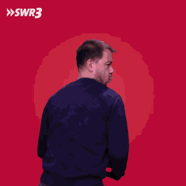 a man in a blue sweater holds a red box in front of a red background with swr3 written on it