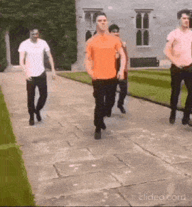 a group of men are walking down a sidewalk . one of the men is wearing an orange shirt .