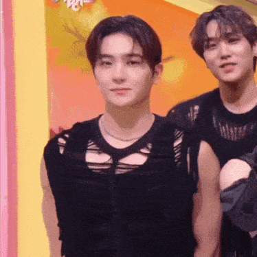 two young men are standing next to each other in front of a yellow wall . one of the men is wearing a black tank top .