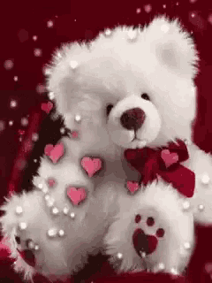 a white teddy bear with hearts and pearls on it 's paws is sitting on a red background .