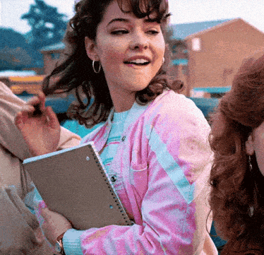 a girl in a pink jacket is holding a notebook and smiling
