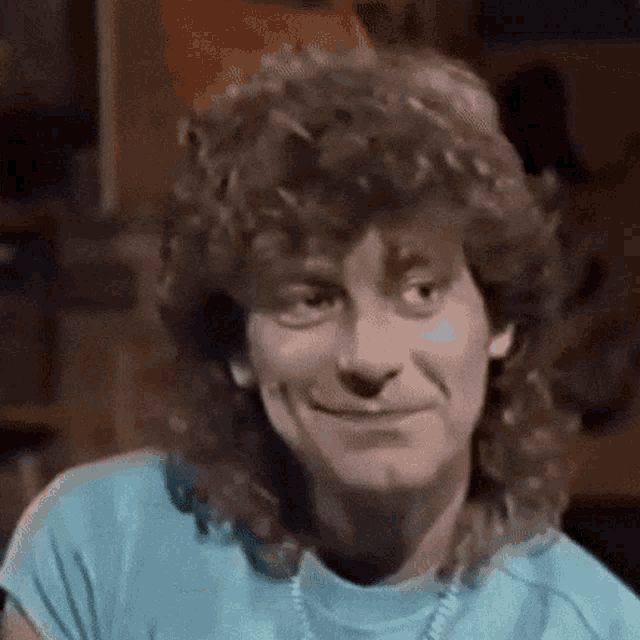 a man with curly hair is smiling and wearing a blue shirt and necklace .